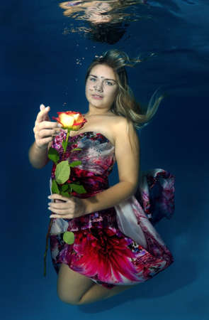 Young beautiful woman in a bright dress with a rose in hands underwaterの写真素材
