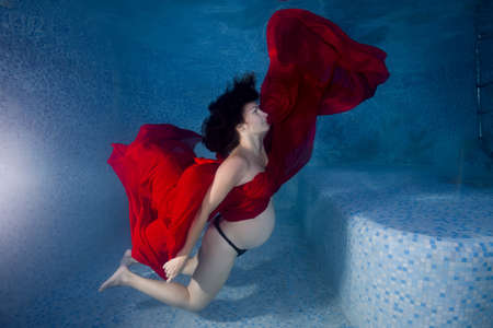Pregnant woman in a red dress posing underwater in the poolの写真素材