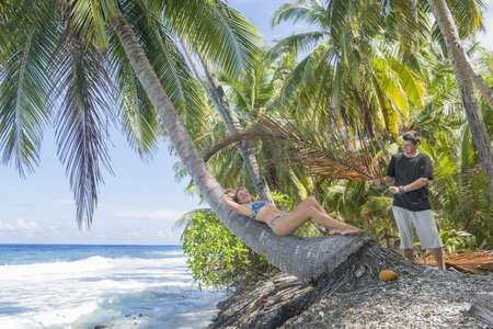 Relaxing woman lies on the coconut palm tree next is a man with a fan on a tropical beach. Young beautiful woman lying on the palm tree on sea shore. Summer vacation concept imageの写真素材