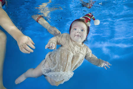 Little girl in a yellow dress and cap swim underwater in the pool, mother holding the child. Healthy family lifestyle and children water sports activity. Child development, disease preventionの写真素材