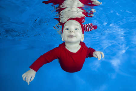 Funny face portrait of little baby boy in red costume swimming and diving underwater with fun in pool on a blue water background. Healthy family lifestyle and children water sports activity.の写真素材