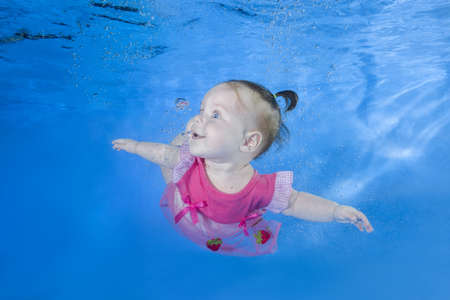 Little baby in pink dress swim underwater. Baby swimming underwater in the pool on a blue water background. Healthy family lifestyle and children water sports activity.の写真素材
