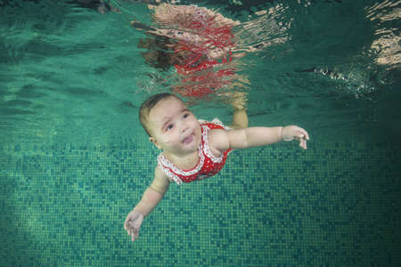A little girl in red swimmiing suit learns to swim underwater in the pool. Baby swimming underwater in the pool. Healthy family lifestyle and children water sports activity. Child development, disease preventionの写真素材