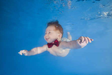 Funny face portrait of baby boy with tongue sticking out swimming and diving underwater with fun in the pool. Healthy family lifestyle and children water sports activity. Child development.の写真素材