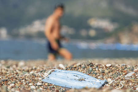 BECICI ANDi BUDVA, BUDVA MUNICIPALITY, MONTENEGRO - JULY 31, 2020: During the COVID-19 pandemic, used medical face masks pollute Discarded used face mask lies on a pebble beach, beachgoers are relaxing by the sea in the background. Becici, Budva Municipality, Montenegroの写真素材