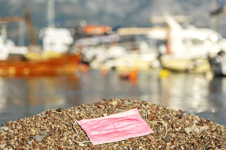 BECICI AND BUDVA, BUDVA MUNICIPALITY, MONTENEGRO - JULY 31, 2020:  Discarded face mask contaminates a beautiful sandy pebble beach, with fishing boats in the background.の写真素材