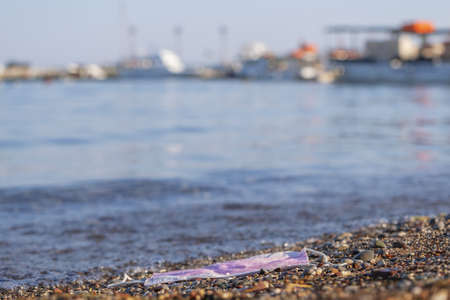 BECICI ANDi BUDVA, BUDVA MUNICIPALITY, MONTENEGRO - JULY 31, 2020: Discarded face mask contaminates a beautiful sandy pebble beach, with fishing boats in the background.の写真素材