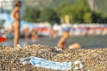 BECICI ANDi BUDVA, BUDVA MUNICIPALITY, MONTENEGRO - JULY 31, 2020: Discarded used face mask lies on a sandy pebble beach, beachgoers are relaxing by the sea in the background.の写真素材