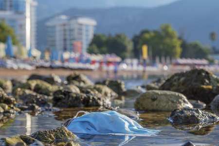 BECICI ANDi BUDVA, BUDVA MUNICIPALITY, MONTENEGRO - JULY 31, 2020:  Discarded used facial mask lies on a pebble beach, in the background a beautiful beach with sun loungers and beach parasols.の写真素材