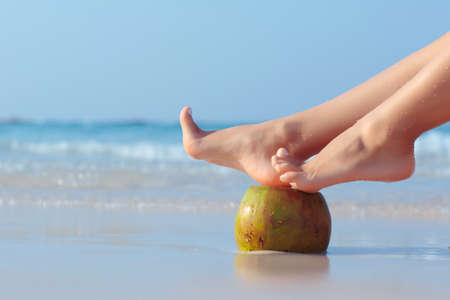Female feet propped on coconut on the beach, blue sea backgroundの写真素材