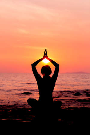 Woman silhouette sitting in lotus position on sunset sea background back litの写真素材