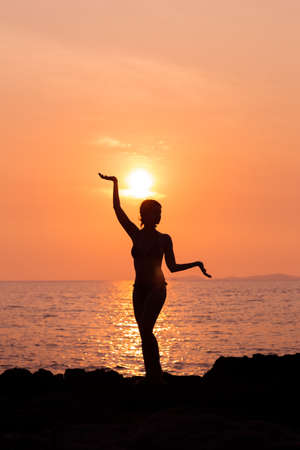 Standing woman silhouette in yoga pose on sunset sea background back litの写真素材
