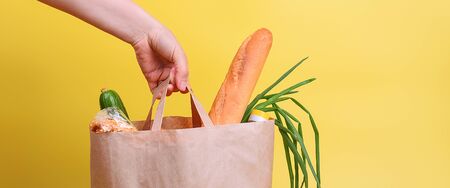 Paper bag with food supplies for the period of quarantine isolation on a yellow background. Delivery, donation, coronavirus. Copyspace. Buckwheat pasta sugar peas canned food tomatoes cucumber bread orange apples eggs ginger.の写真素材