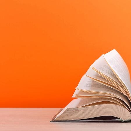 A large, thick hardcover book lies on a light-wood table. The book is open, the leaves of the page are fanned out, and the back is a orange background with space for an inscription, text, or logo.の写真素材