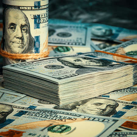 A roll of dollars with coins on the background of scattered one hundred dollar bills in blue light.