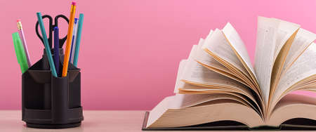 A large thick book with the pages spread out like a fan and a stand with pens, pencils and scissors on the table on a pink background.の写真素材
