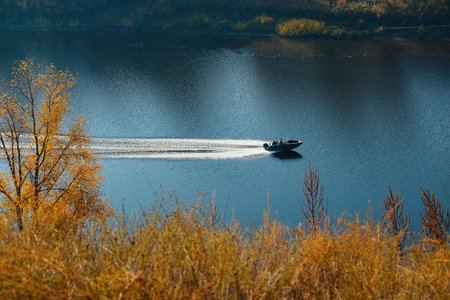 The motor boat moves in the middle of the blue river flowing between the hills with autumn forest....
