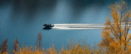 The motor boat moves in the middle of the blue river flowing between the hills with autumn forest. Beautiful scenery.の写真素材
