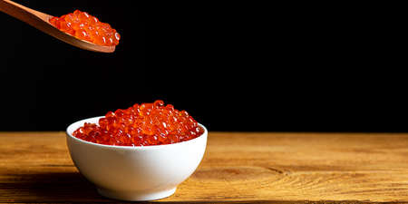 Red caviar in a wooden cup on a black background with a spoon. A large pile of bright caviar. Fresh delicious caviar. A place for advertising, logo, label, layout, mockup.の写真素材