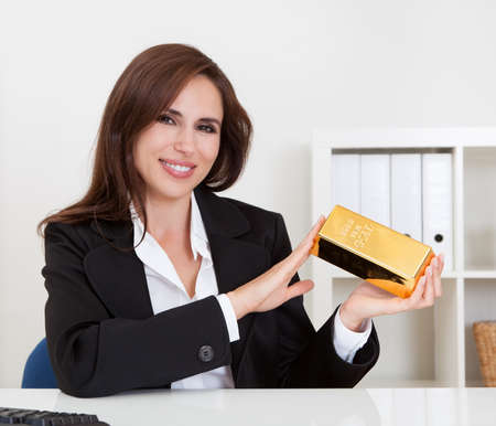 Portrait of a beautiful young businesswoman holding gold bar at office deskの写真素材