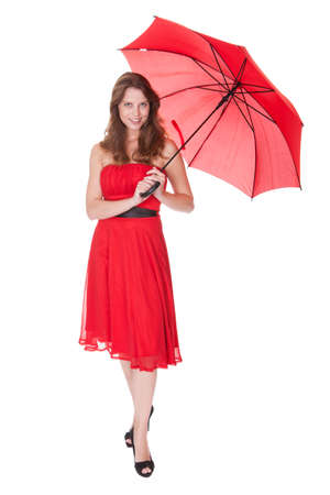 Beautiful elegant woman in a trendy red dress posing under an open red umbrella isolated on whiteの写真素材