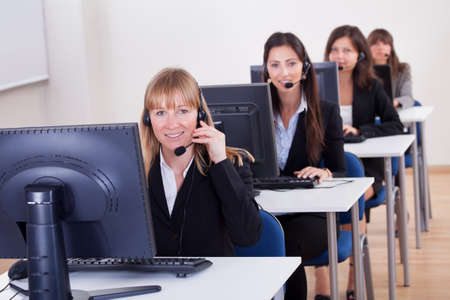 Row of attractive young telephonists seated at computers wearing headsets and microphones in a call centre or client services help deskの写真素材