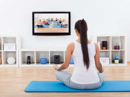 Rear view of a spiritual young woman sitting on a mat meditating at home while watchingの写真素材