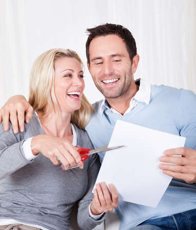 Laughing woman about to cut up a document poised with the scissors at the ready while being watched by her husbandの写真素材