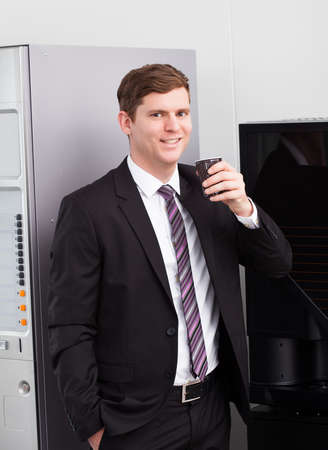Portrait of happy businessman drinking coffee standing near vending machineの写真素材