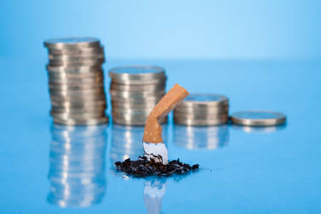 Single Cigarette Butt With Ash And Coin Stack On Blue Backgroundの写真素材