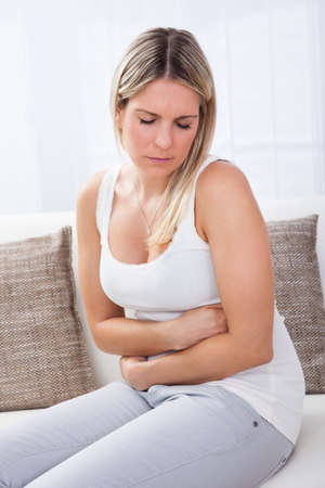 Portrait of woman with stomach ache sitting sofaの写真素材