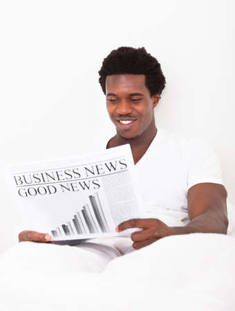 Portrait Of Casual Young African Man Reading Newspaper Sitting On Bedの写真素材