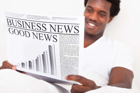 Portrait Of Casual Young African Man Reading Newspaper Sitting On Bedの写真素材