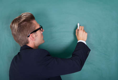 Portrait Of Young Male Professor Writing On Chalkboard In Classroom