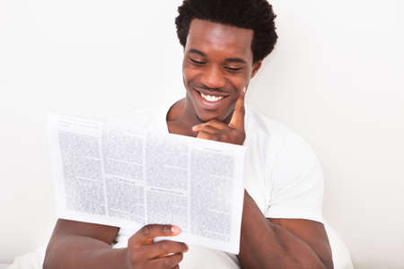 Portrait Of Casual Young African Man Reading Newspaper Sitting On Bedの写真素材