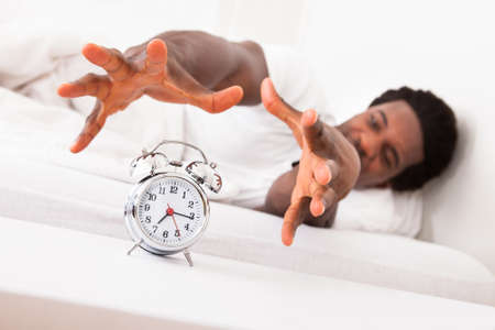 Frustrated Man Trying To Catch Is Alarm Clock While Relaxing On His Bedの写真素材