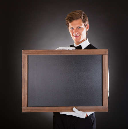 Happy Young Male Waiter Holding Chalkboard Over Black Background
