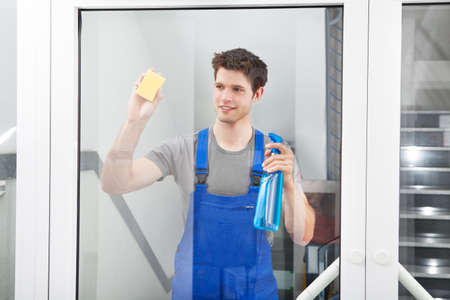 Young Male Cleaner Cleaning The Door Glass With Sponge And Spray Bottleの写真素材