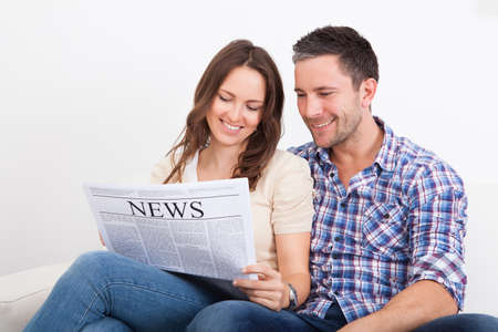 Portrait Of A Happy Young Couple Sitting On Couch Reading Newspaperの写真素材