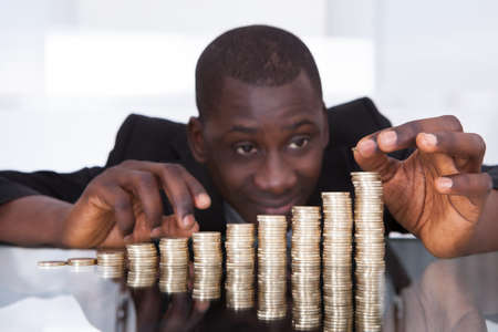 Portrait Of A Young African Businessman Stacking Coins In A Row On Desk