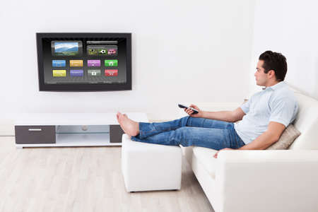 Young Man At Home Applying Setting Of Television Sitting On Couch