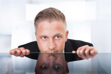 Businessman peering furtively over the top of his desk as he spies on a colleague or tries to remain...