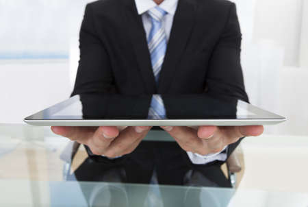 Businessman holding a tablet computer in his hands offering it to the viewer with a smile as he sits at his desk in the officeの写真素材