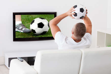 Excited soccer fan watching a game on television holding a soccer ball above his head as he sits on a comfortable sofa in his living roomの写真素材