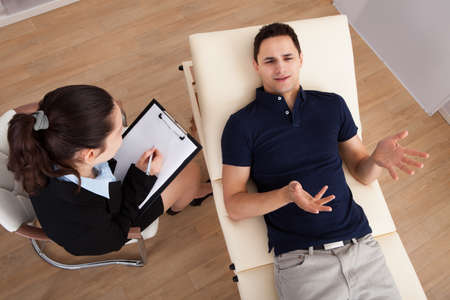 High angle view of male patient communicating while psychologist writing notes on clipboard in clinicの写真素材