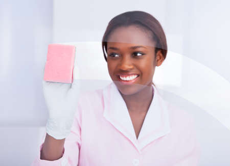 Happy African American female housekeeper cleaning glass in hotelの写真素材