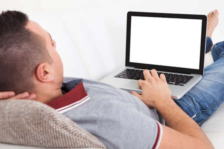 Young man using laptop while lying on sofa at homeの写真素材
