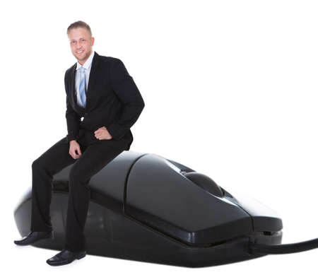 Smiling confident businessman in a suit sitting on a wired computer mouse with a scrolling wheel in on online communication and e-commerce concept  on whiteの写真素材