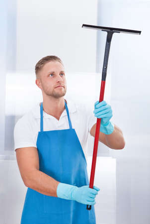 Male janitor using a squeegee to clean a window in an office wearing an apron and gloves as he works