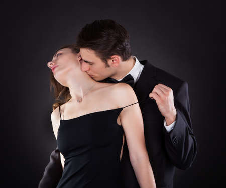 Young man in suit kissing woman on neck while removing dress strap from her shoulder isolated over black backgroundの写真素材
